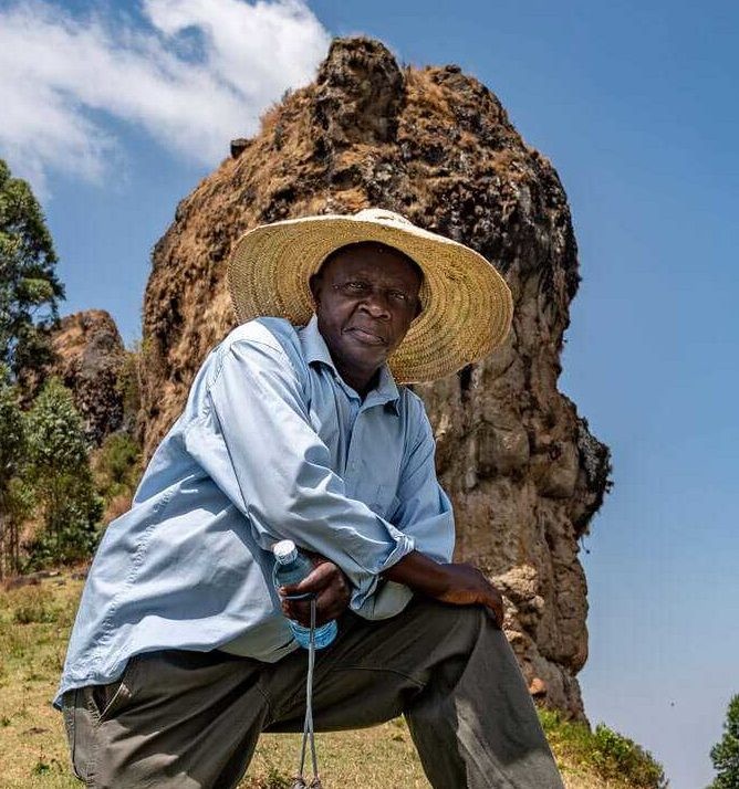 Sie sehen ein Bild von Nirmord dem Vorstand der Bio Fairtrade Kaffee Bauern in Uganda, Afrika. Er trägt ein Hemd und einen großen Hut und steht auf der Spitze des Berges Mount Elgon. Dort bauen die Farmer feinsten Bio Fairtrade Kaffee an.